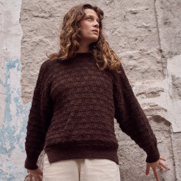 Woman wearing a brown knitted sweater against a textured wall.