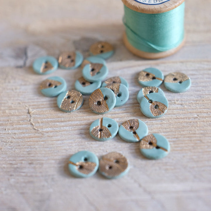 Turquoise buttons with gold centers on a wooden surface next to a spool of thread.
