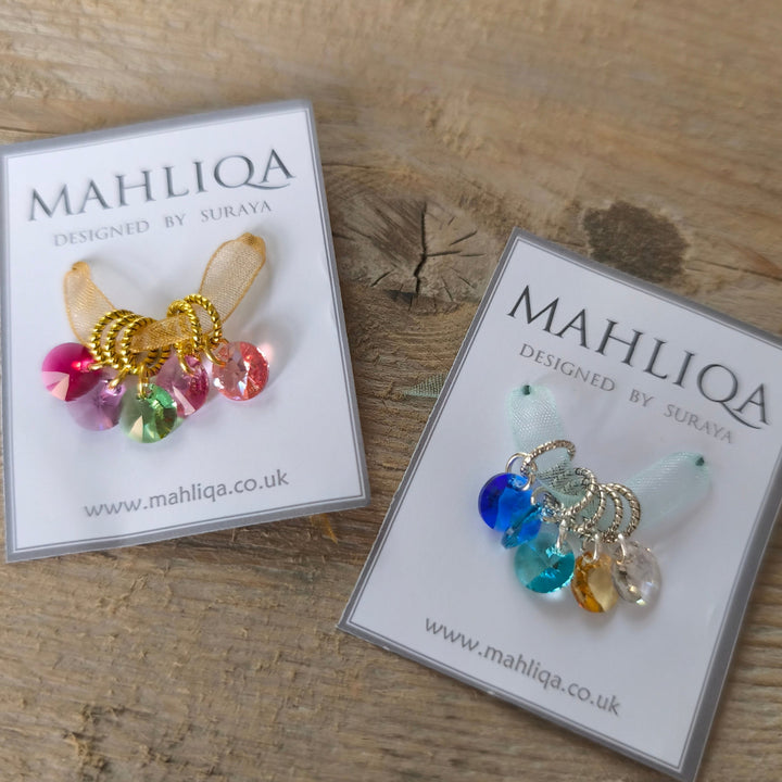 Two packs of colorful stitch markers on display with 'MAHLIQA' packaging on a wooden surface.