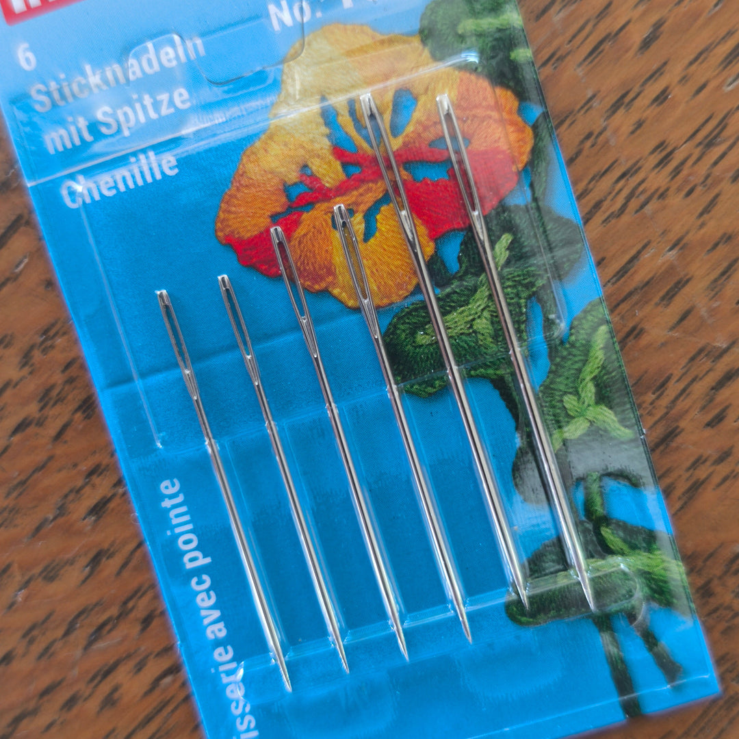 Six metal embroidery needles in a blue plastic blister pack with a floral illustration and German text, placed on a wooden surface.