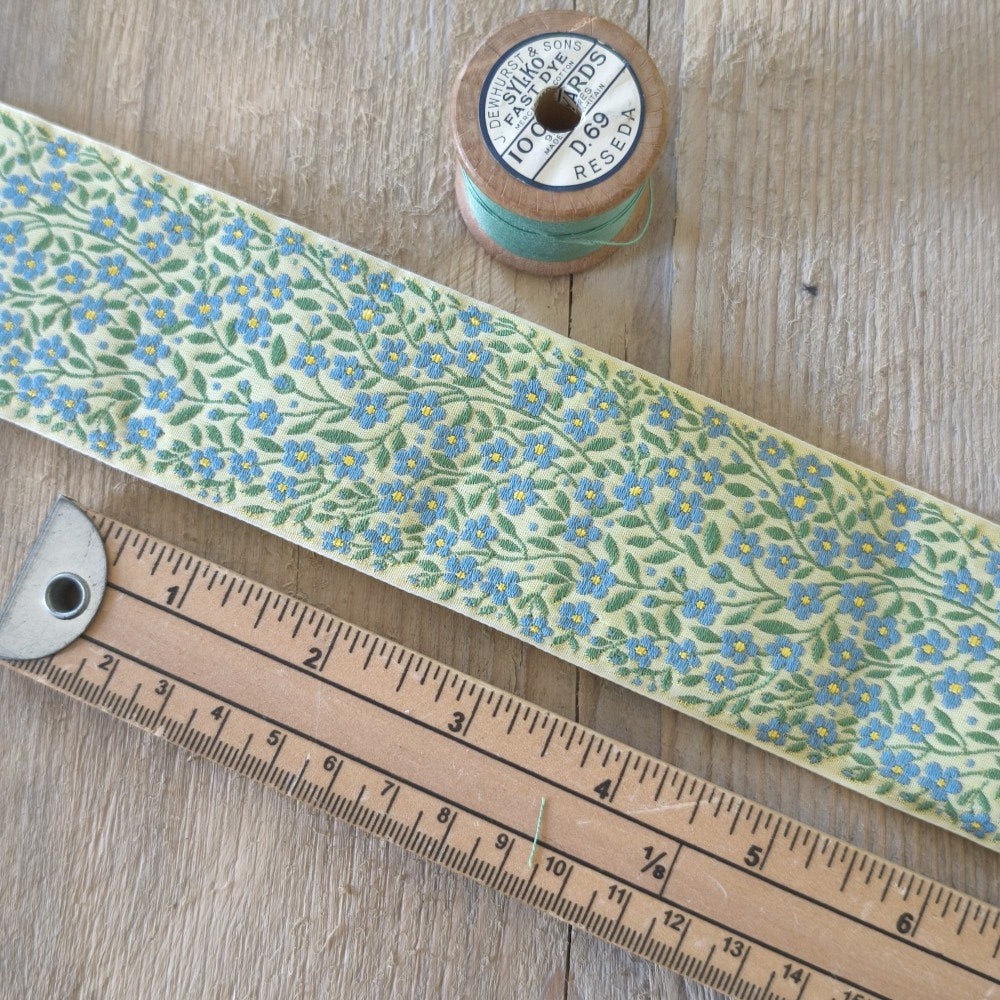 A close-up of a patterned ribbon or strip laid diagonally on a wooden surface, shown with a wooden ruler and a spool of thread for scale and context. The product itself has a pale yellow-green base with a repeating floral print in blue and green.