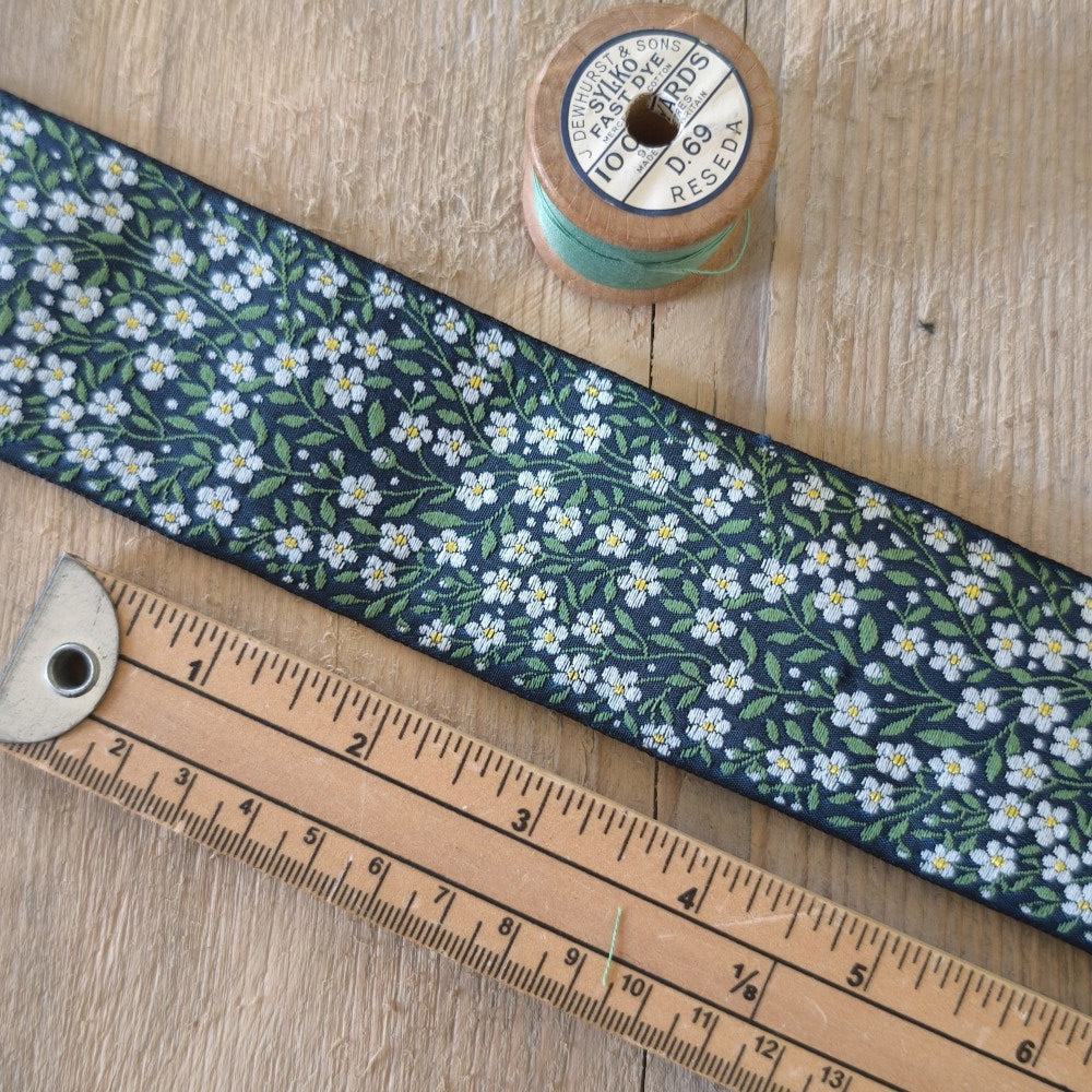 A close-up of a fabric ribbon with a navy blue background and a repeating white and yellow forget-me-not flower pattern with green leaves. The ribbon is laid diagonally on a light wood surface next to a wooden ruler and a spool of light blue thread.
