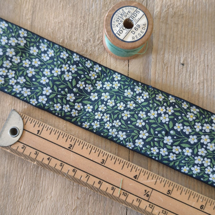 A close-up of a fabric ribbon with a navy blue background and a repeating white and yellow forget-me-not flower pattern with green leaves. The ribbon is laid diagonally on a light wood surface next to a wooden ruler and a spool of light blue thread.