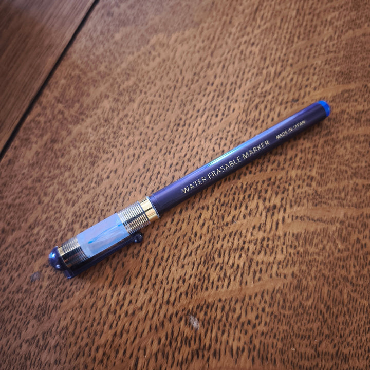 A blue and gold water erasable marker pen resting diagonally on a textured brown wooden surface.