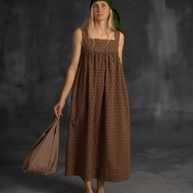 Woman wearing a brown patterned dress and green headscarf, holding a woven bag against a dark background.