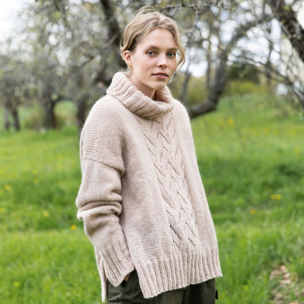 Woman wearing a beige knitted sweater in a grassy field with trees in the background