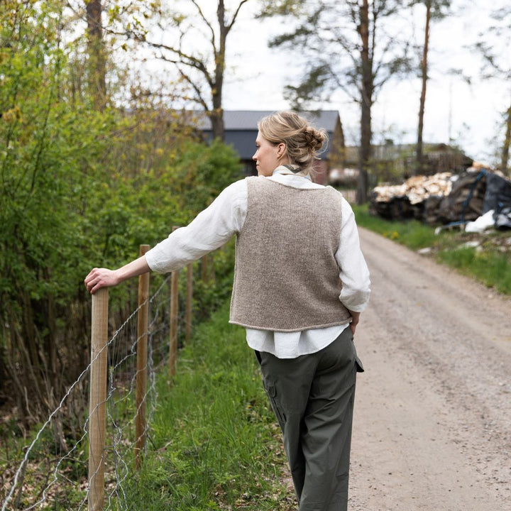 Person wearing a natural knitted vest on a path next to a fence with trees and buildings in the background