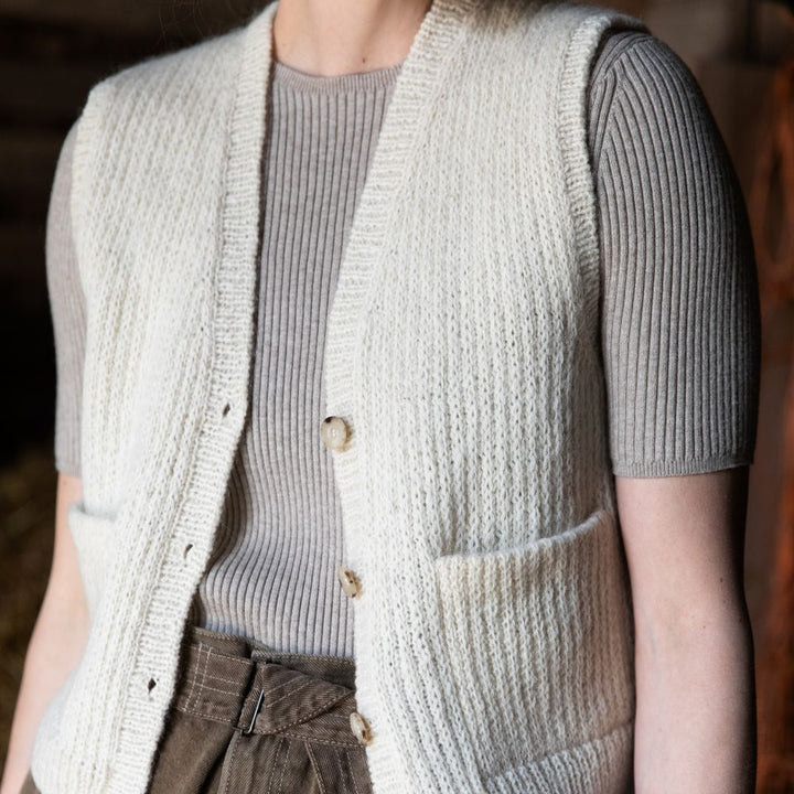 Person wearing a cream knitted vest over a gray shirt with brown pants.