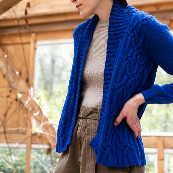 Woman wearing a bright blue textured cardigan, beige sweater, and brown pants indoors.