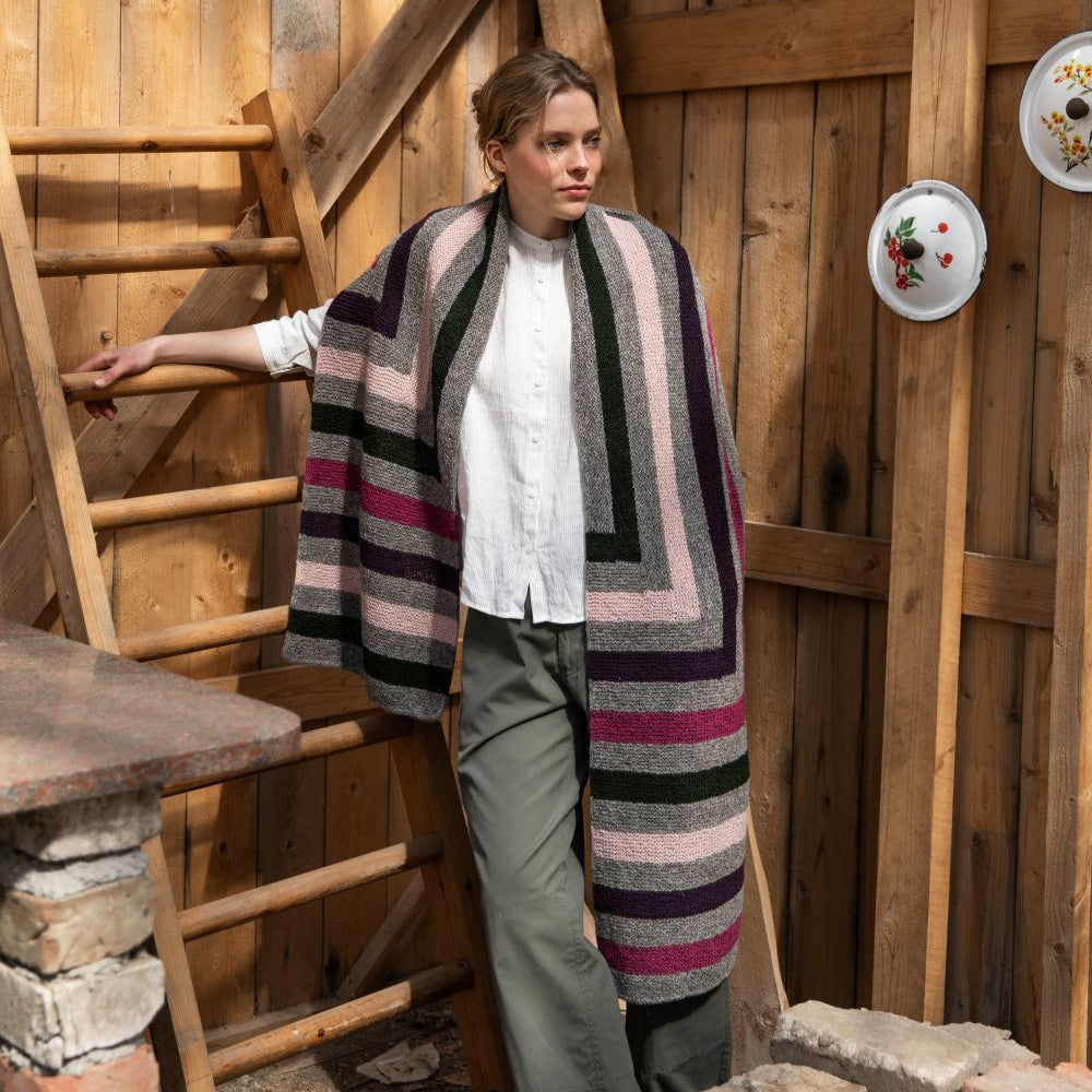 Person wrapped in a long striped knitted wrap standing in a rustic wooden interior.