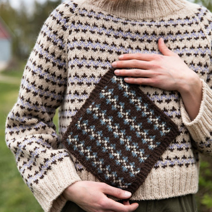 Person wearing a patterned sweater holding a knitted swatch outdoors.